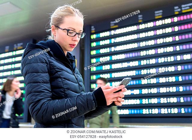 Woman in international airport looking at smart phone app information and flight information board, checking her flight detailes