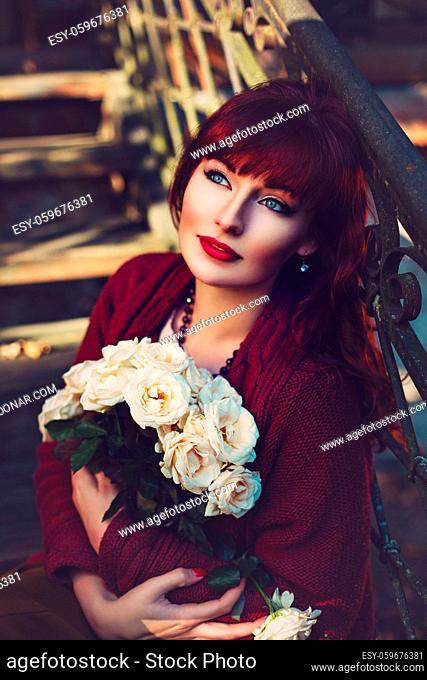 Beautiful young woman in long skirt and wool red scarf sitting on old house stairs holding flowers. Early fall. Outside shot. Ambient light