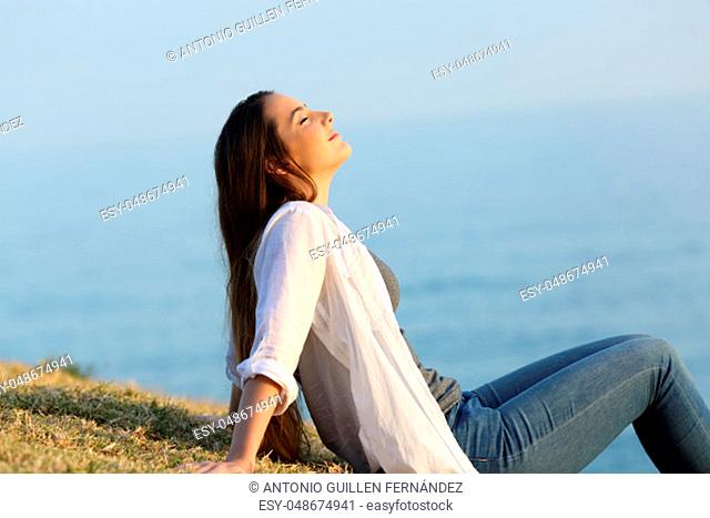 Side view portrait of a relaxed woman breathing fresh air sitting on the grass with the sea and sky in the background