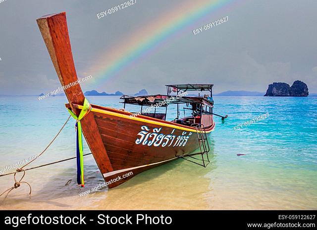 Longtail boat in crystal clear blue waters waiting for passengers on beautiful white beach in Ko Mok, Thailand after rain with a beautiful rainbow