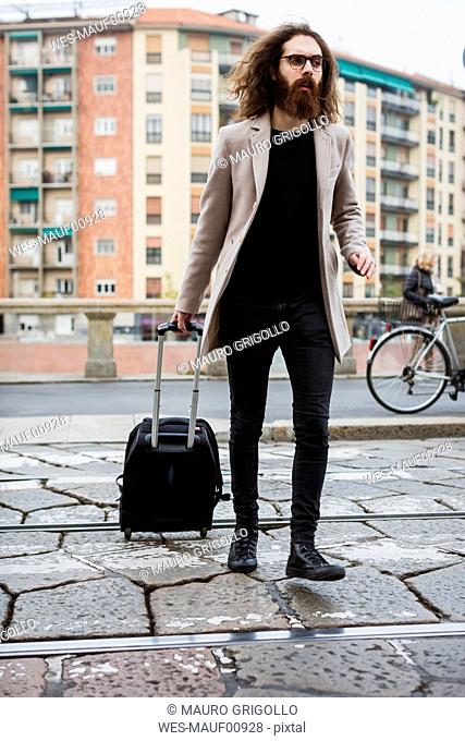 Stylish young man with suitcase crossing tramway