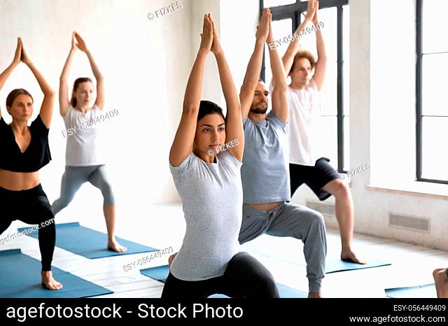 Young diverse people practicing yoga, doing Warrior one exercise, beautiful Indian woman instructor standing in Virabhadrasana pose at group lesson