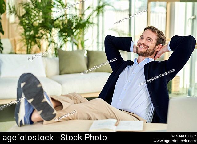 Cheerful male freelancer looking away sitting with feet up on table in home office