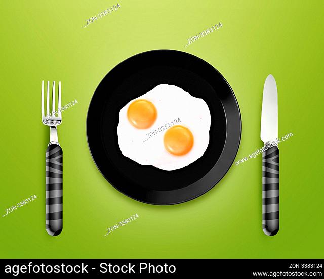 top view of two fried eggs on black Plate between silver knife and fork on gray background