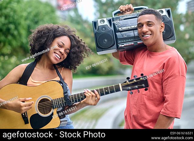 Best time. Happy dark-skinned girl with guitar and guy holding record-playerr on shoulder smiling at camera standing in park