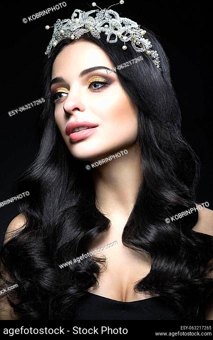 Beautiful dark-haired woman with a crown of precious stones, curls and evening makeup. Beauty face. Picture taken in the studio on a black background