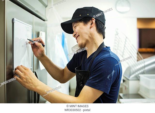 Man working in a bakery, wearing baseball cap and apron, writing note on small whiteboard, using phone and smiling