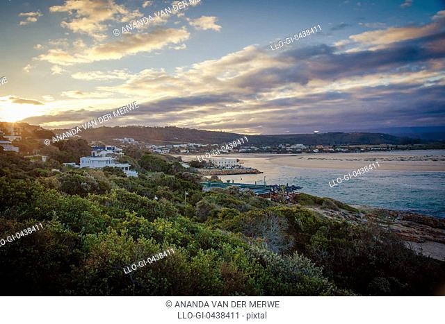 The sun sets behind a mountainous backdrop in a spectacular sky overlooking the beautiful homes and restaurants of the town of Plettenberg Bay and the flow of...