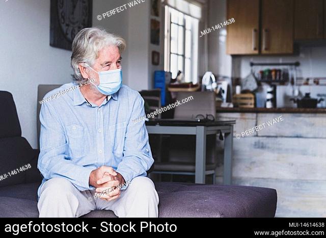 portrait of one old and mature man sitting on the sofa wearing mask to prevent coronavirus indoors - lockdown lifestyle at home