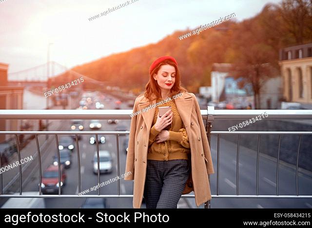 Portrait of stylish young woman wearing autumn coat and red beret outdoors. Tender young woman warming up with cup of coffee on the street female fashion...