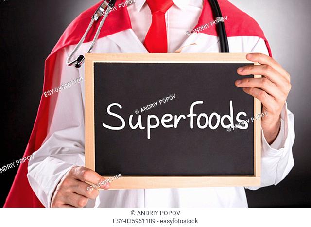 Close-up Of Superhero Doctor Holding Small Chalkboard With The Text Superfoods