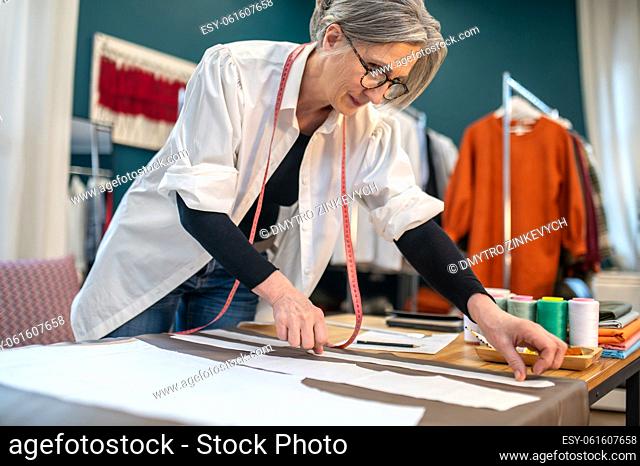 Accuracy. Focused middle aged woman in glasses standing over table applying paper pattern to material in atelier
