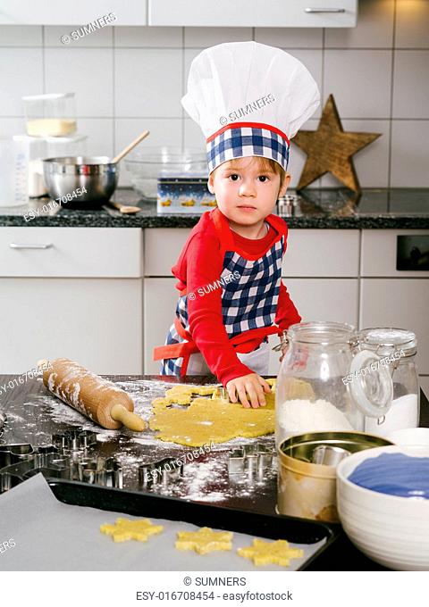 Photo of an adorable boy in a chef hat and apron making snowflake cookies in the kitchen