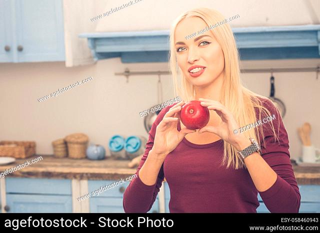 Toned picture of lady posing with apple in kitchen at home. Happy beautiful blond woman showing her healthy lifestyle. Woman smiling for camera