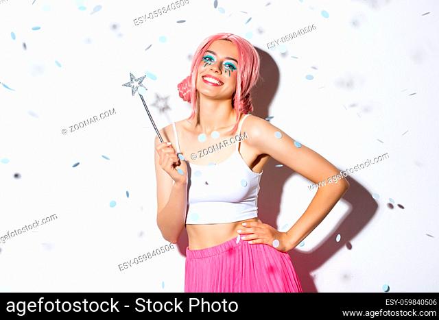 Image of beautiful happy girl in pink wig, celebrating halloween in fairy costume, holding magic wand and smiling while confetti floating in air