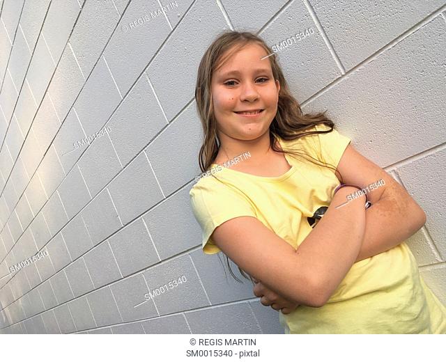 young girl leaning against a white brick wall and looking at the camera while crossing her arms
