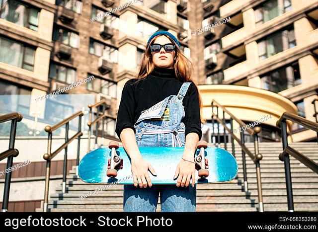 Stylishly dressed woman in blue denim jumpsuit posing with skateboard. Street photo. Portrait of girl holding skateboard. Lifestyle, youth concept