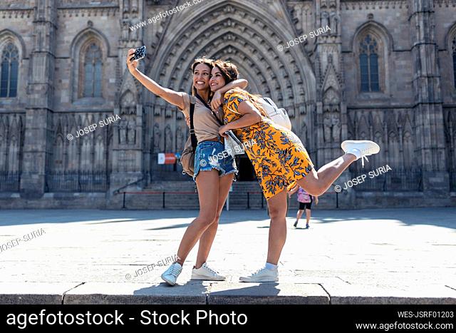 Woman embracing friend while taking selfie through mobile phone standing at Barcelona Cathedral Square in Barcelona, Catalonia, Spain
