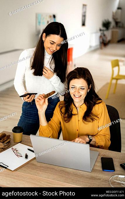 Smiling mother having cookie while using laptop by daughter holding mobile phone at desk in home office