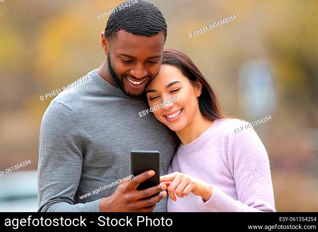 Happy interracial couple checking smart phone outdoors