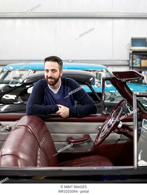 A portrait of a Caucasian male leaving on the side of his classic car convertible in a repair shop