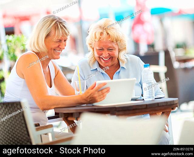 Senior women in outdoor cafe on sunny summer day. They looking at pad screen and laughing. Enjoyable time with friend and tablet PC