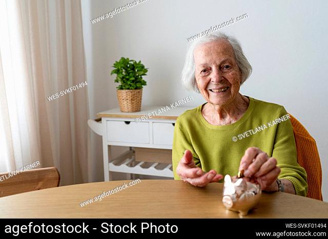 Smiling woman putting coin in piggy bank on table