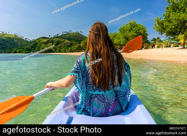 Rear view of a young woman with long brown chestnut hair paddling a canoe along the shore of an idyllic island during vacation in Indonesia