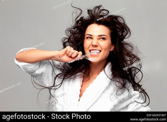 A brunette woman in a white terry robe with disheveled hair brushes her teeth in the morning after sleep. Concept of personal hygiene, morning time