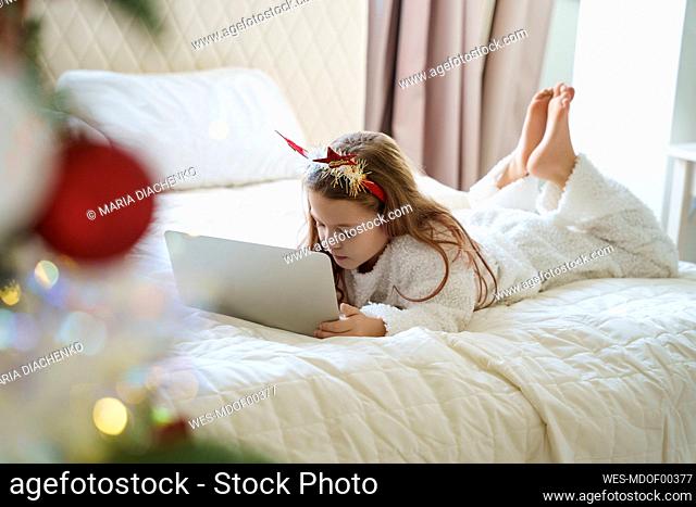Girl wearing Christmas headband lying on bed and using laptop at home