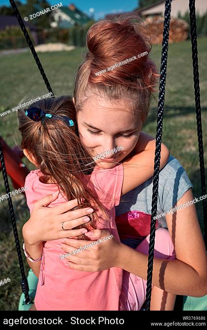 Teenage girl hugging her younger sister in a home playground in a backyard. Happy smiling sisters having fun on a swing together on summer day