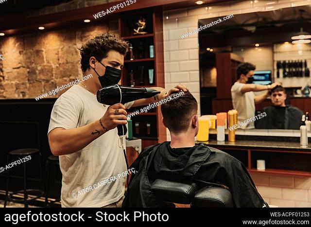 Male barber using hair dryer for young man in salon during COVID-19