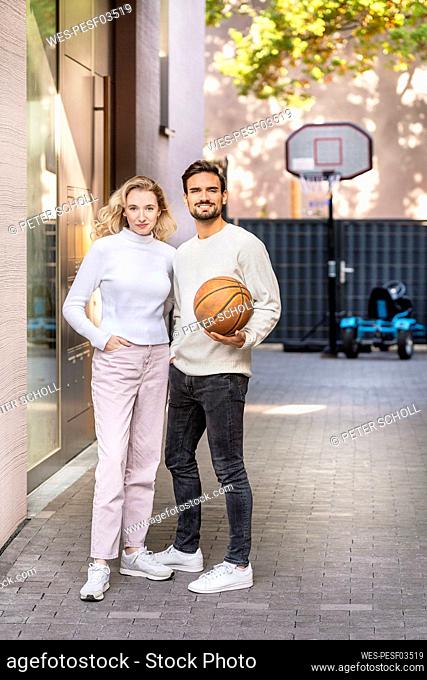 Young man holding basketball standing with girlfriend on footpath