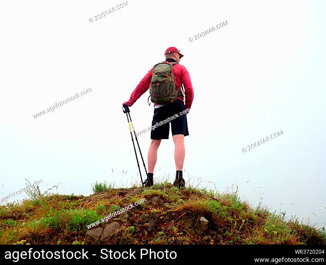 Man in cloud of fog. Hiker in pink black sportswear and poles stand on mountain peak rock. Around heavy mist. Poor visibility