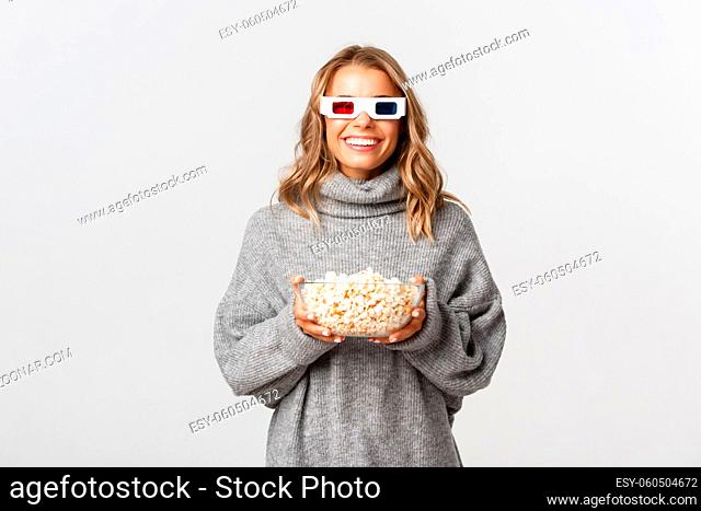 Image of beautiful young woman in grey sweater, watching movies in 3d glasses and eating popcorn, smiling happy at camera, standing over white background