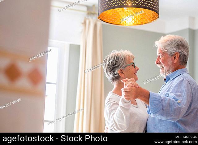 Cheerful senior couple dancing and smiling looking at each other. Elderly happy couple celebrating by dancing in living room