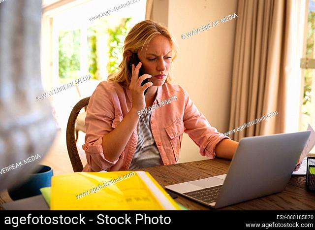 Caucasian woman sitting at table working in living room, using laptop and talking on smartphone