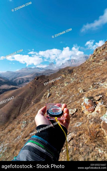 A man's hand holds a pocket magnetic compass for navigation against the background of a rocky slope and epic rocks under a blue sky and white clouds