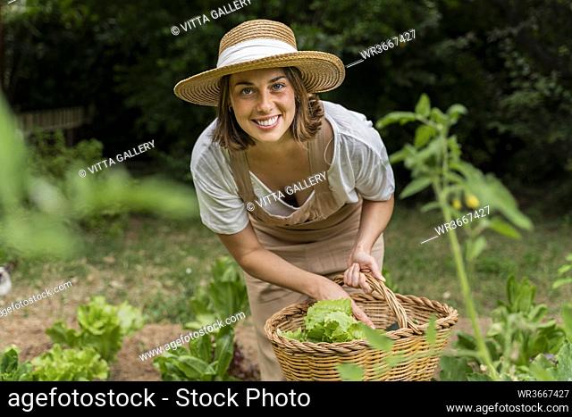 Smiling young woman wearing hat collecting vegetables in wicker basket at yard