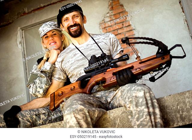Smiling male and female military persons with crossbow weapon are sitting and looking at camera