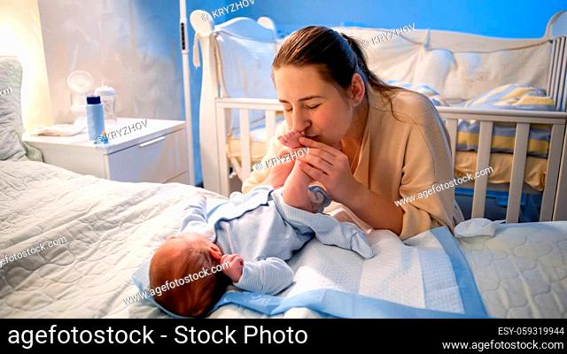 Happy smiling young mother looking at her newborn baby boy lying on bed at night and kissing his feet. Concept of family happiness and parenthood