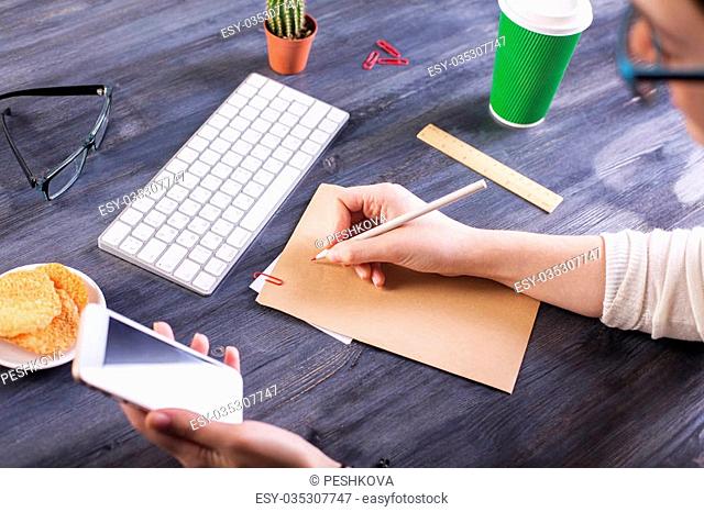 Top view of woman's hands copying information from smart phone to brown paper on wooden desktop with keyboard, cookies, glasses