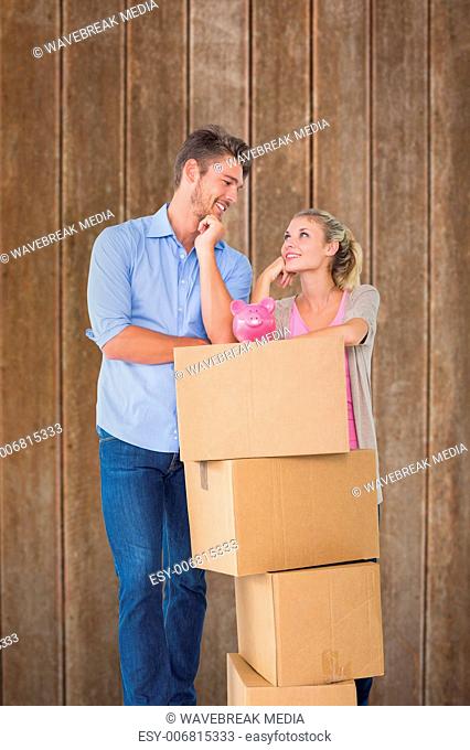 Composite image of attractive young couple leaning on boxes with piggy bank