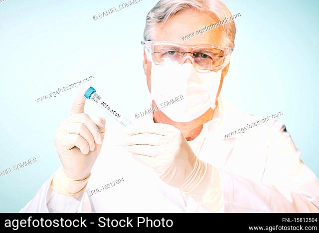 Grey-haired laboratory technician with protective goggles and face mask looking at test tube with Cornoravirus sample