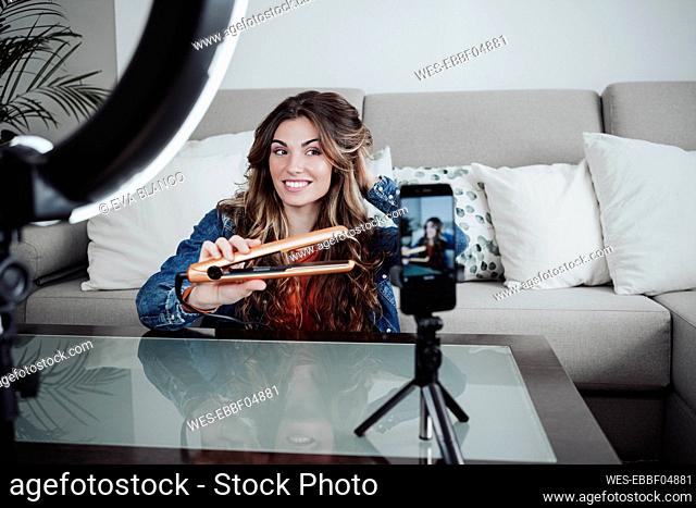 Smiling hairstylist curling hair with machine and vlogging through smart phone at home studio