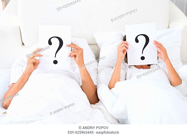 Couple Lying On Bed Holding Question Mark Over Faces