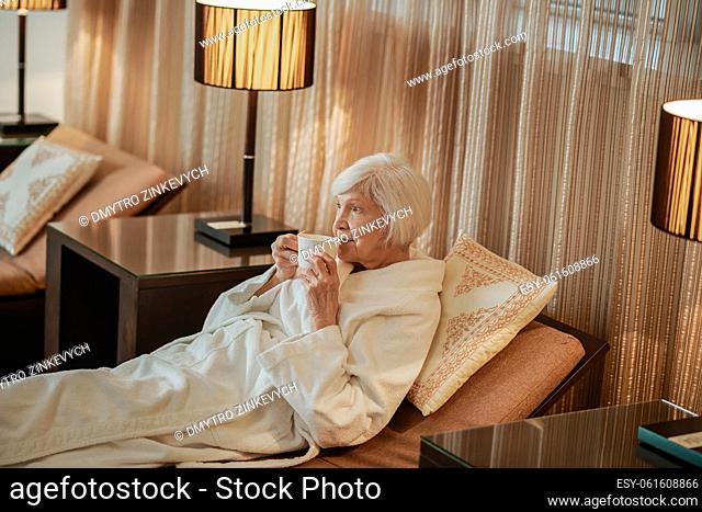 Morning coffee. A senior gray-haired woman lying in bed and having morning coffee