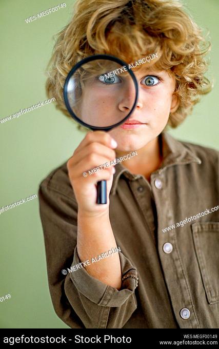 Serious boy looking through magnifying glass against green background