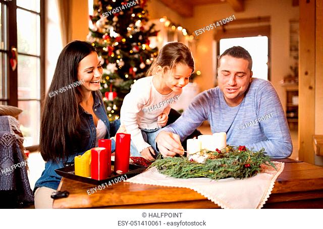 Beautiful young family with their daughter at home lighting candles on advent wreath. Illuminated Christmas tree behind them