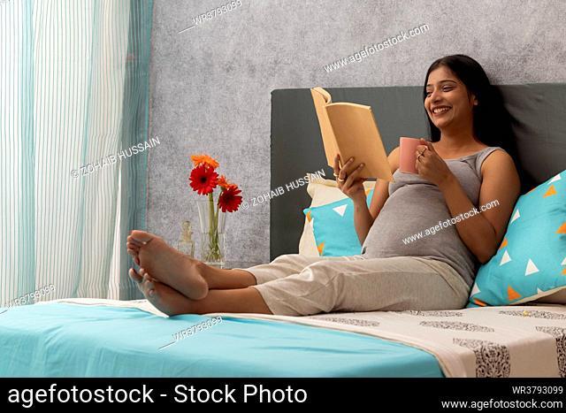 Pregnant woman drinking hot drink and reading a book while relaxing on bed at home